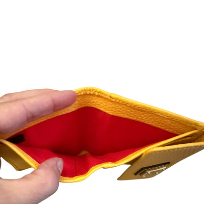 A close-up of a hand holding open a small yellow leather wallet, showing a bright red interior lining and a gold-tone metal clasp on a plain white background.