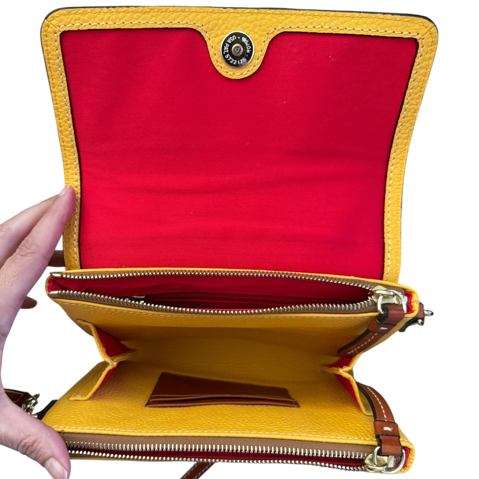 A yellow pebbled leather crossbody bag is shown open, revealing a bright red interior lining, gold-tone zippers, card slots, and a button closure. A hand is holding the left side of the bag against a plain white background.