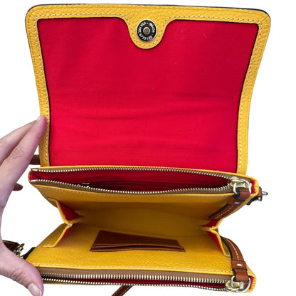 A yellow pebbled leather crossbody bag is shown open, revealing a bright red interior lining, gold-tone zippers, card slots, and a button closure. A hand is holding the left side of the bag against a plain white background.