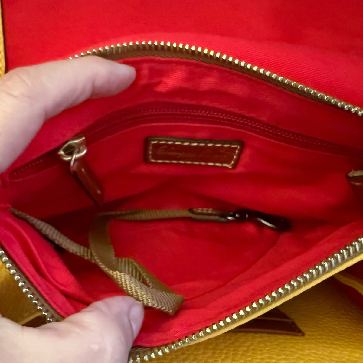 Close-up of the interior of a handbag showing a bright red fabric lining, gold-tone zipper trim, a small stitched brand pacth, and part of a tan woven strap. A hand is holding the opening to show the inside.