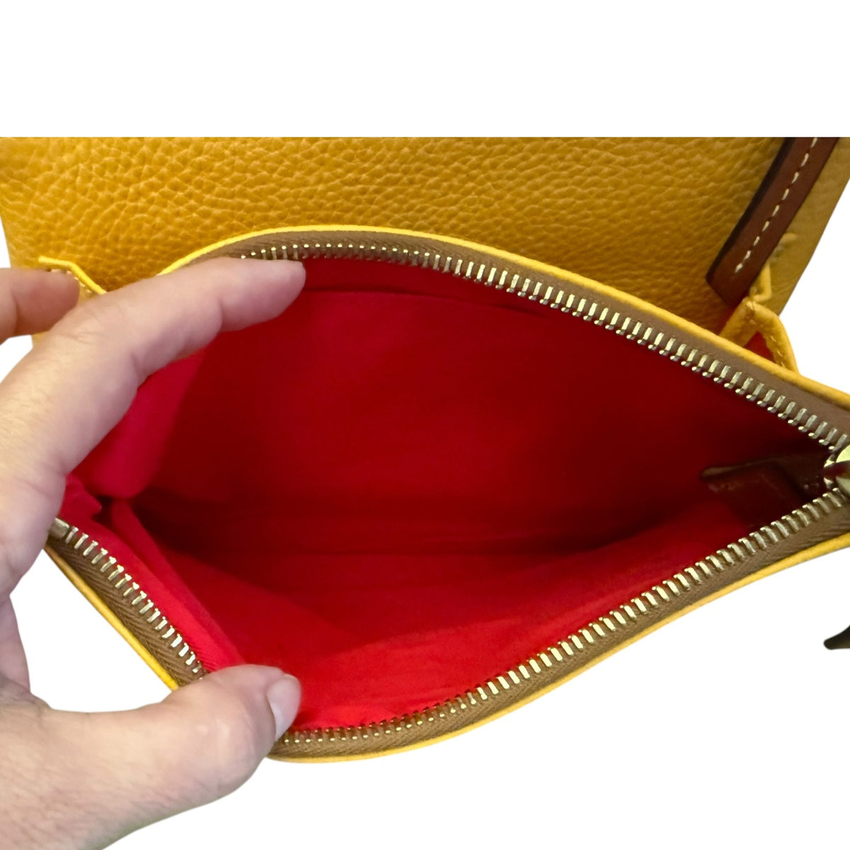 A close-up of a yellow pebbled leather bag being opened by a hand, showing a bright red interior lining and a gold-tone zipper.
