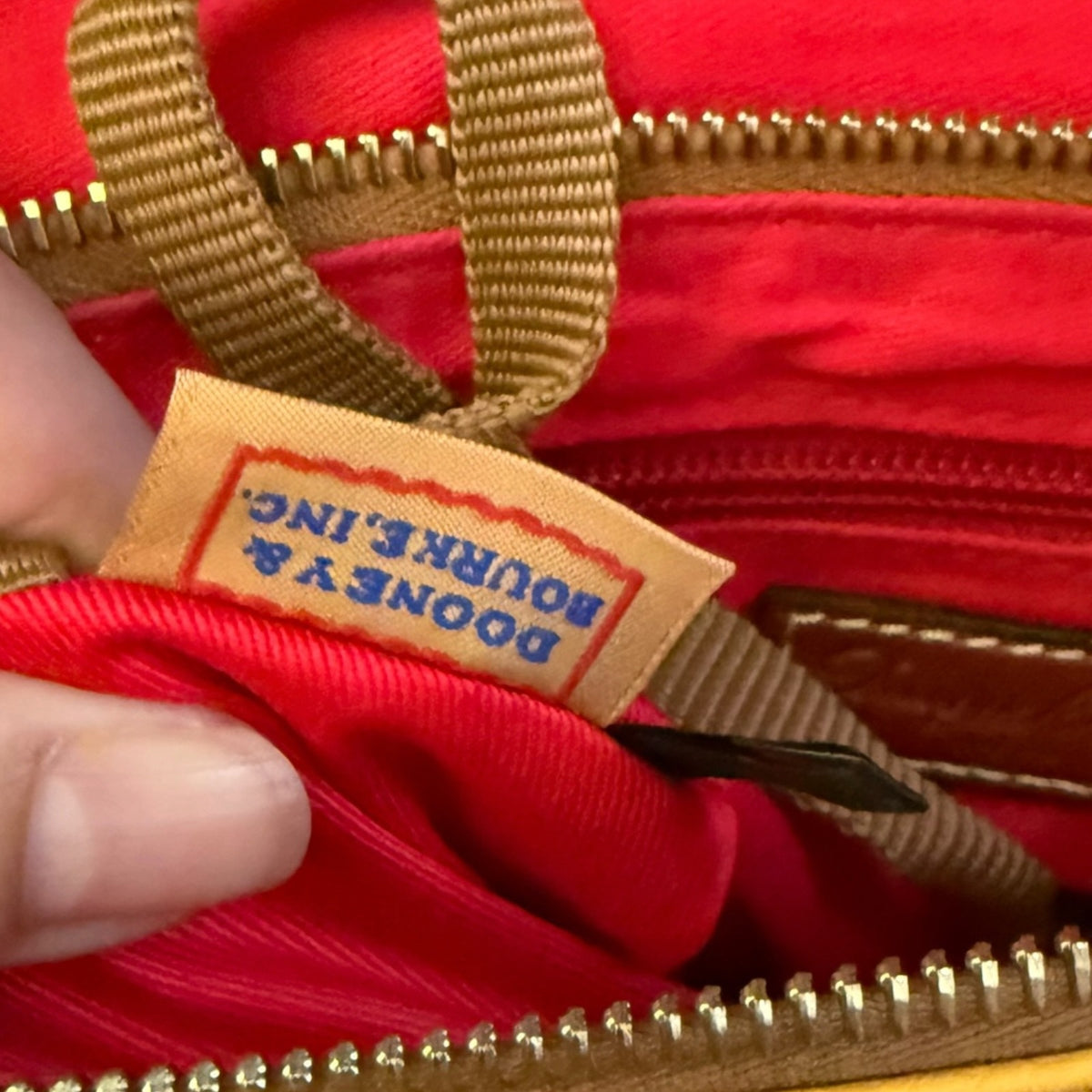 Close-up of a red interior of a bag with tan straps and gold-tone zippers, showing a sewn label that reads DOONEY & BOURKE, with a finger holding the opening open.