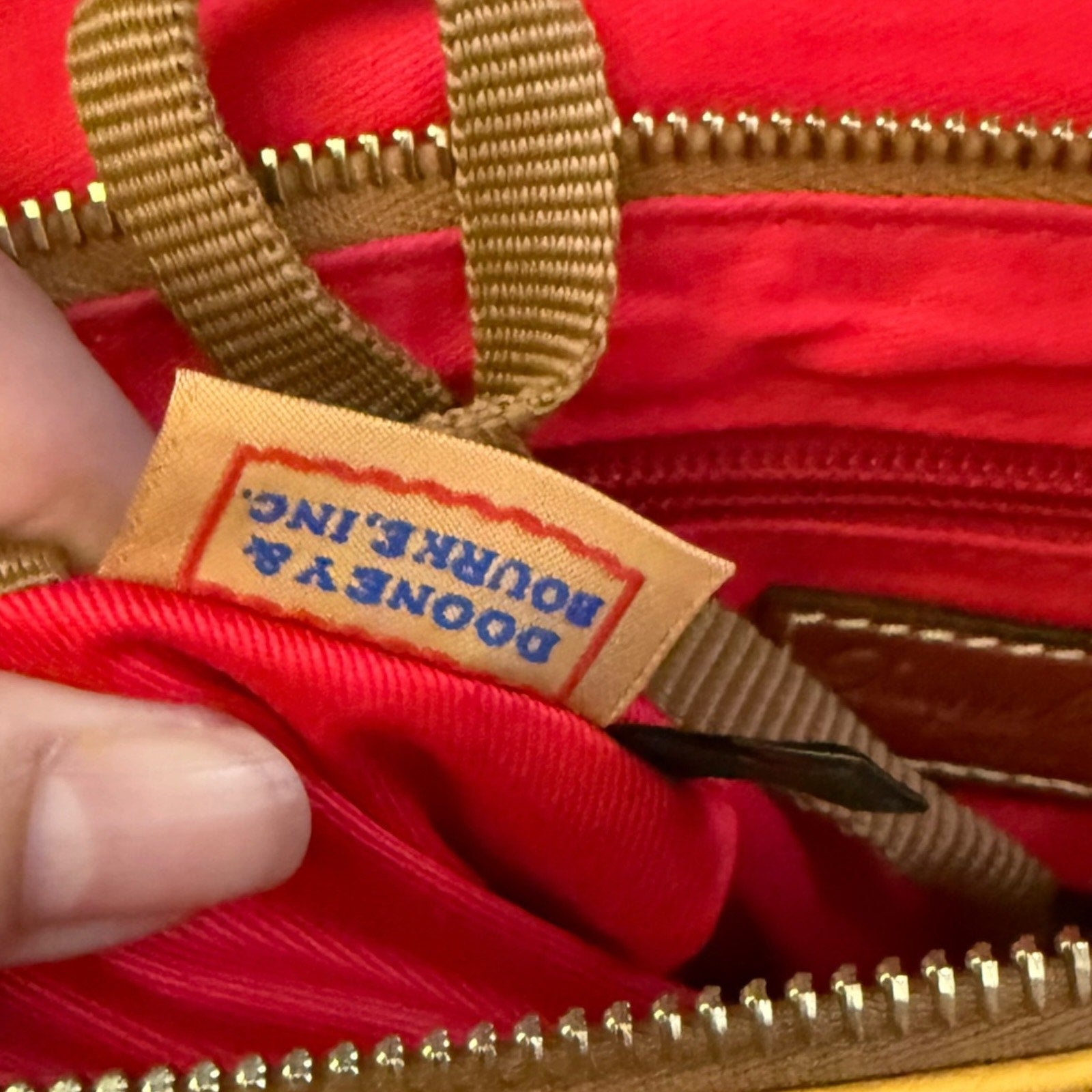 Close-up of a red interior of a bag with tan straps and gold-tone zippers, showing a sewn label that reads DOONEY & BOURKE, with a finger holding the opening open.