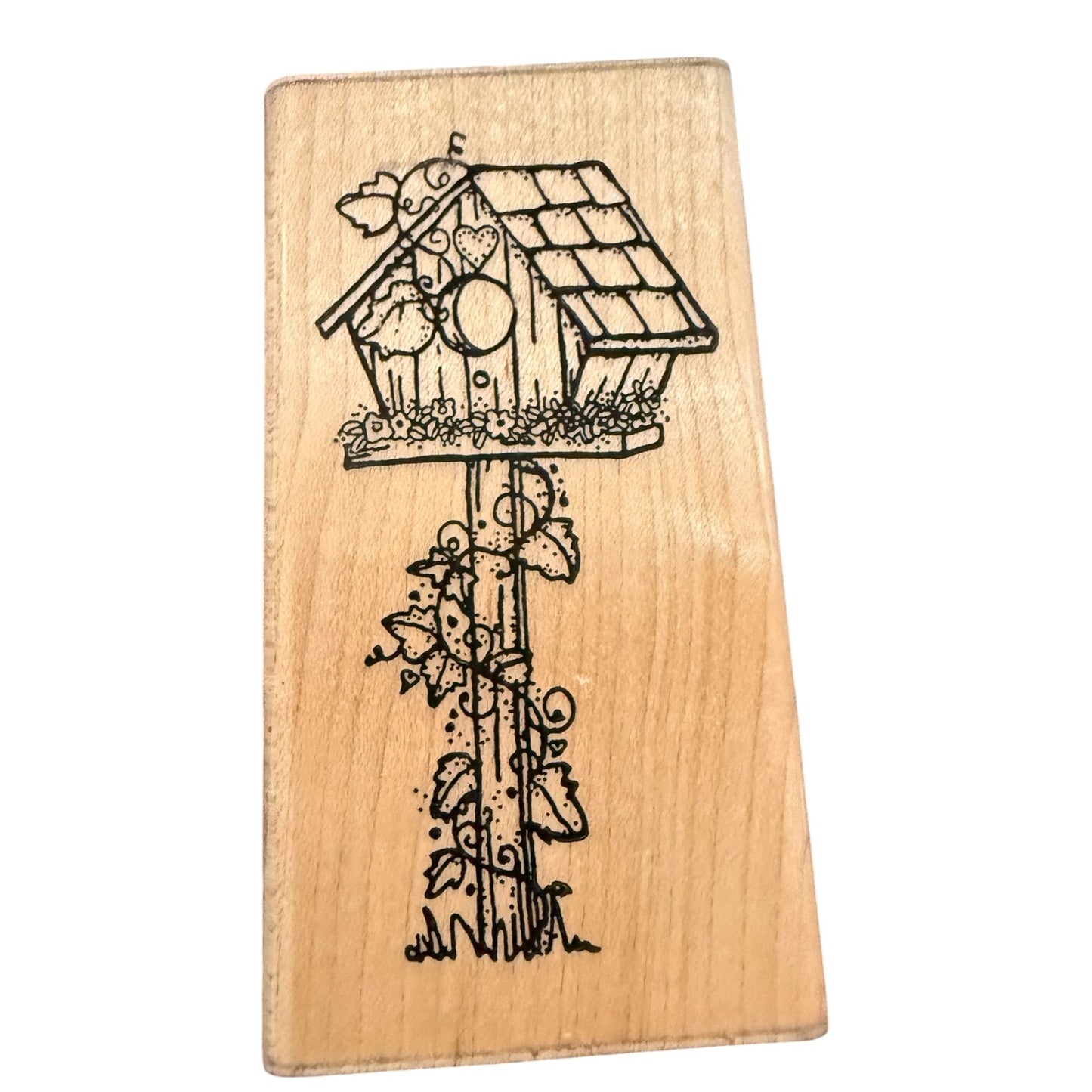 A wood stamp block featuring  birdhouse with a bird perched on it, climbing vines, and small flowers, shown against a plain white background.