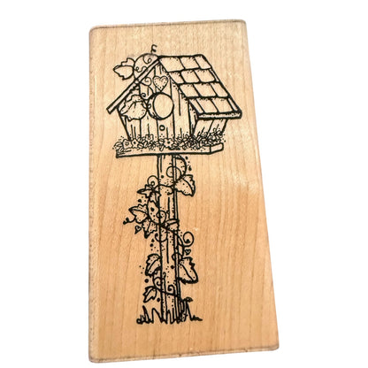 A wood stamp block featuring  birdhouse with a bird perched on it, climbing vines, and small flowers, shown against a plain white background.
