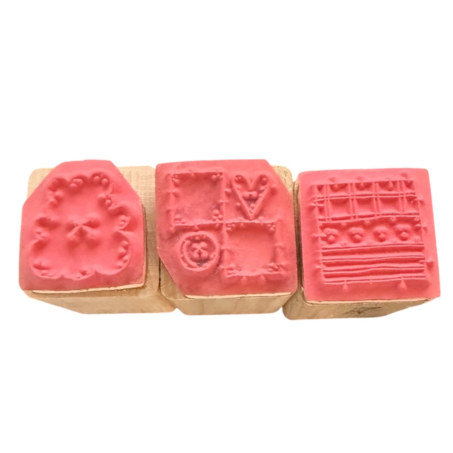 Three wooden rubber stamps with pink-red stamps showing floral, geometric, and striped patterns, arranged side by side on a white background.