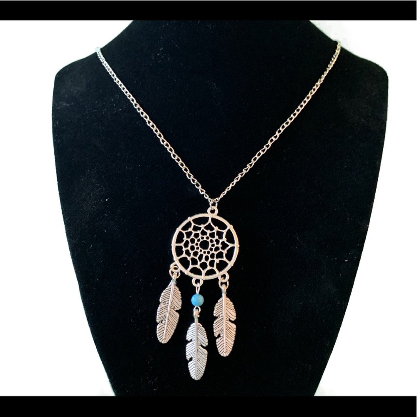 A dreamcatcher-style pendant necklace displayed on a black jewelry bust. The necklace has a silver-tone chain and a circular web design with three hanging feather charms in silver accented by a small turquoise bead.