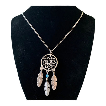 A silver dreamcatcher necklace displayed on a black velvet jewelry bust. The necklace features a silver-toned chain, a circular webbed dreamcatcher pendant, and three hanging feather charms, with one feather accented by a small blue bead.