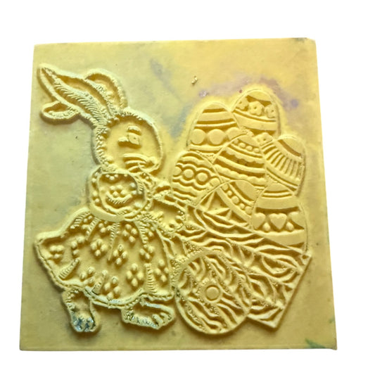 A square yellowish rubber stamp featuring an Easter bunny and patterned Easter eggs, shown on a plain white background.