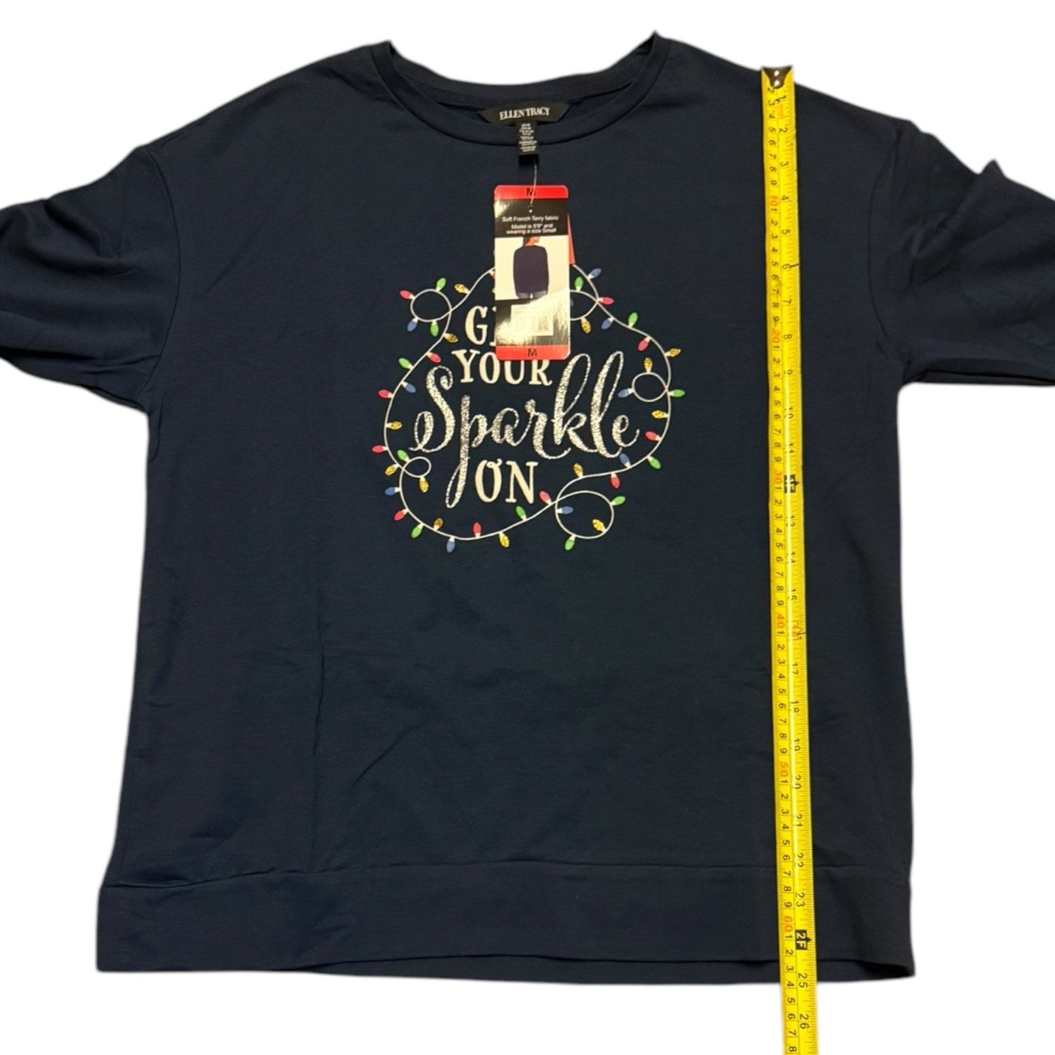 Blue shirt with 'Turn Your Sparkle On' text and Christmas lights design, with a measuring tape on a white background.