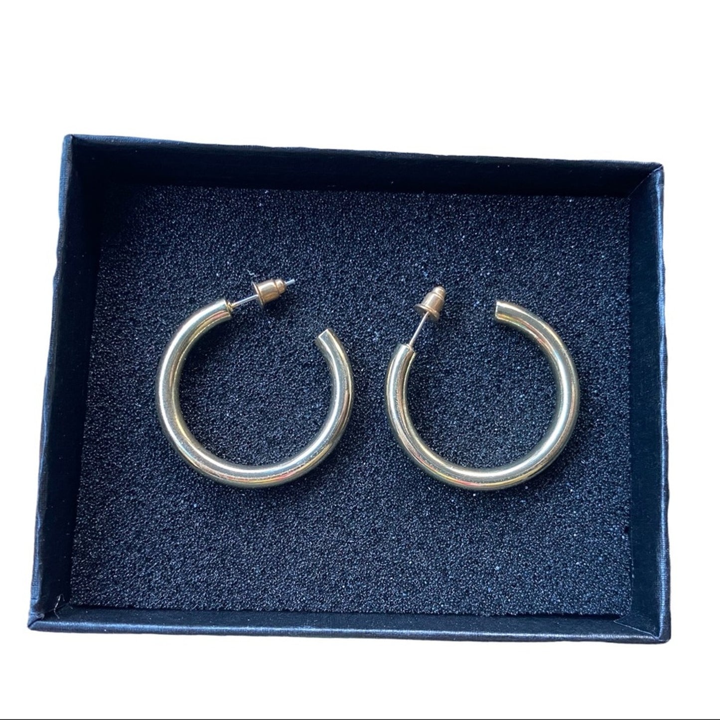 A pair of polished gold-tone hoop earrings displayed inside an open  presentation box with a dark blue  insert.