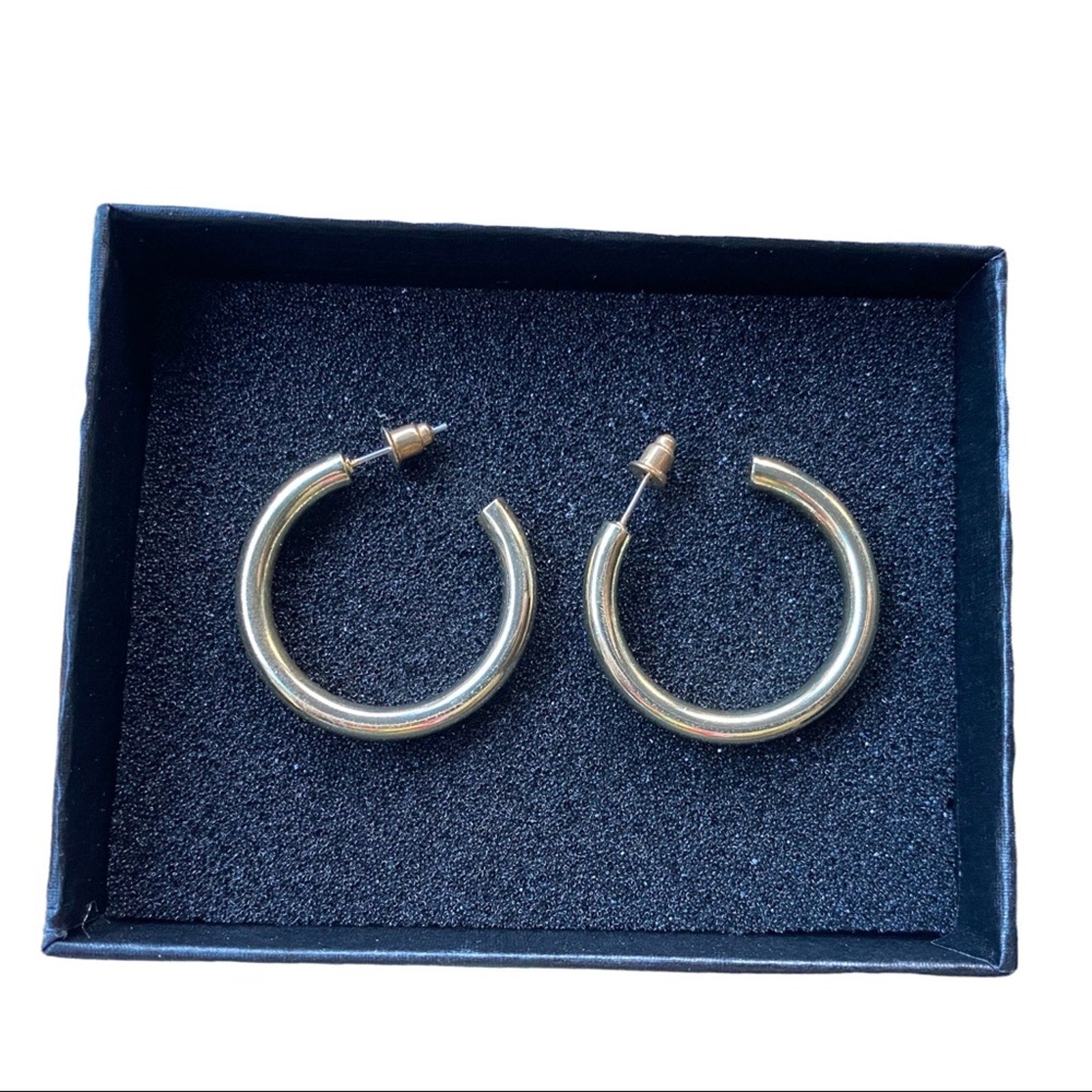 A pair of polished gold-tone hoop earrings displayed inside an open  presentation box with a dark blue  insert.