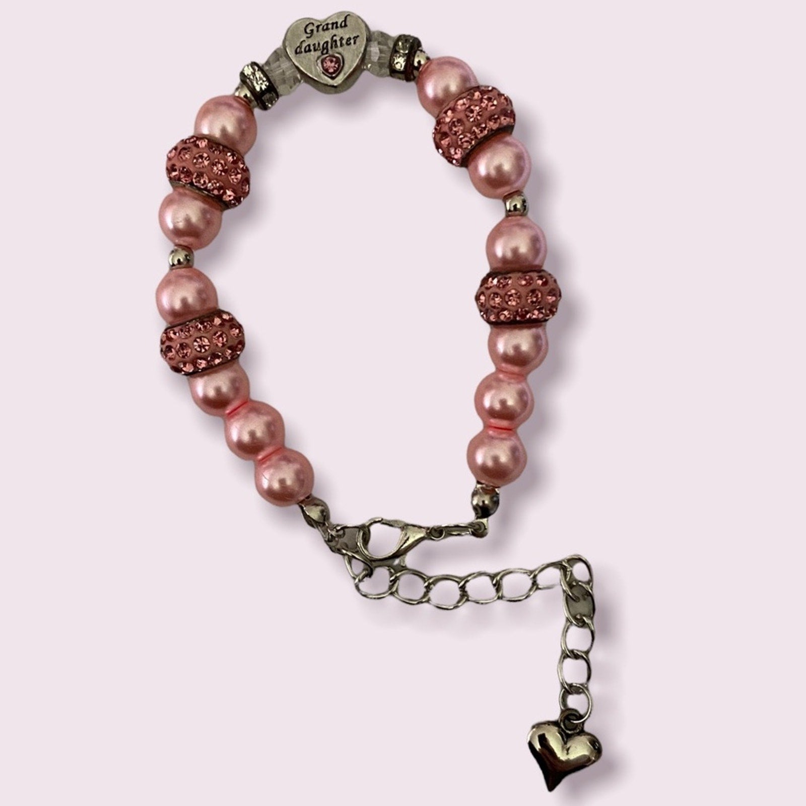 A beaded bracelet with pink faux pearl beads, rose gold rhinestone-encrusted beads, and antique bronze-toned metal accents, including a heart-shaped charm engraved with 'Grand daughter' and a dangling heart charm, shown on a light pink background.
