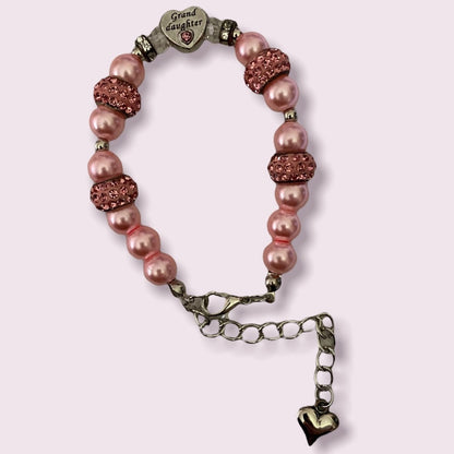 A beaded bracelet with pink faux pearl beads, rose gold rhinestone-encrusted beads, and antique bronze-toned metal accents, including a heart-shaped charm engraved with 'Grand daughter' and a dangling heart charm, shown on a light pink background.