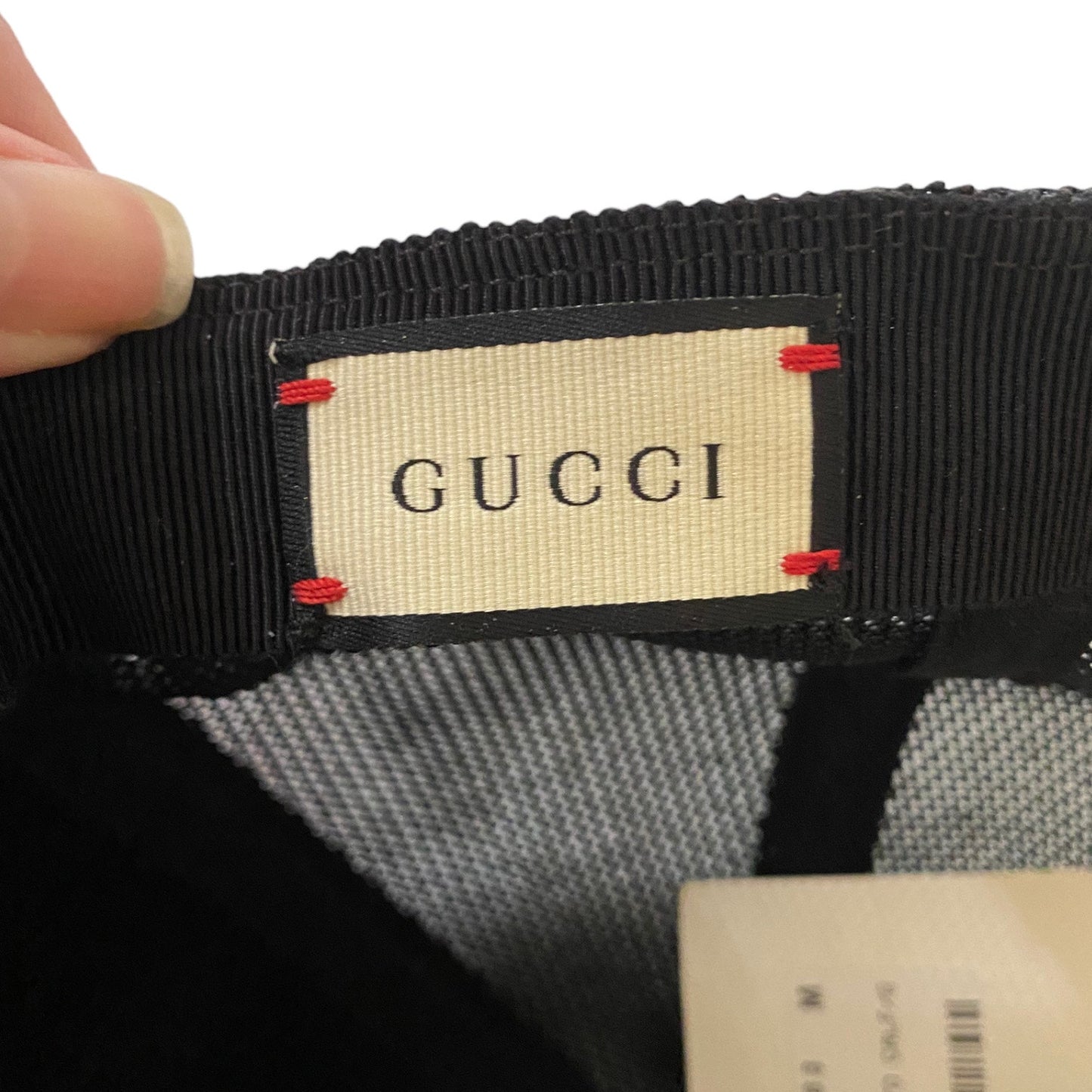 Close-up of a Gucci hat's interior label area, showing a beige Gucci brand patch stitched onto a black ribbed band with small red accent stitches, held by a finger against a white background.