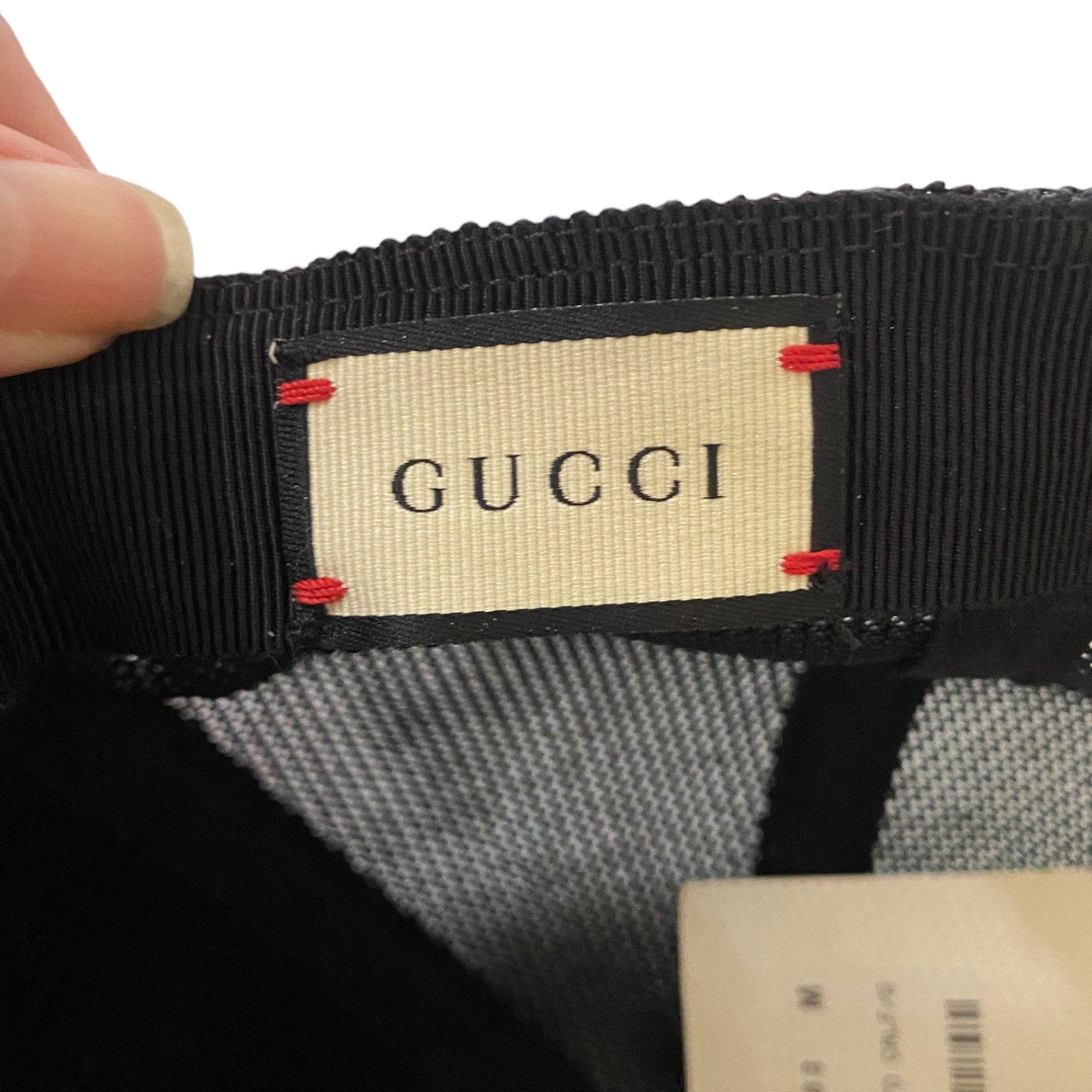 Close-up of a Gucci hat's interior label area, showing a beige Gucci brand patch stitched onto a black ribbed band with small red accent stitches, held by a finger against a white background.