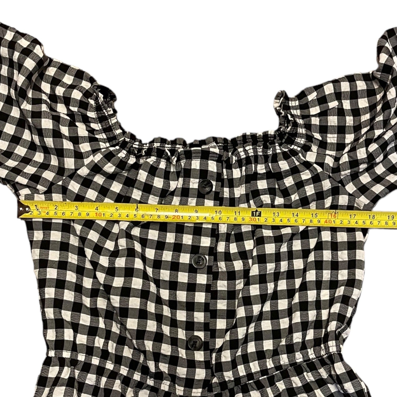 Black and white gingham check top with  a yellow measuring tape stretched horizontally across the chest to show the width.