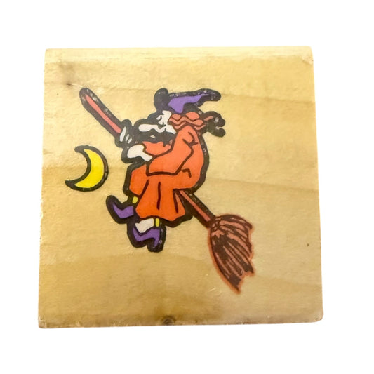 A square wooden rubber stamp featuring an illustration of a witch riding a broomstick with a yellow crescent moon, set against a plain white background.
