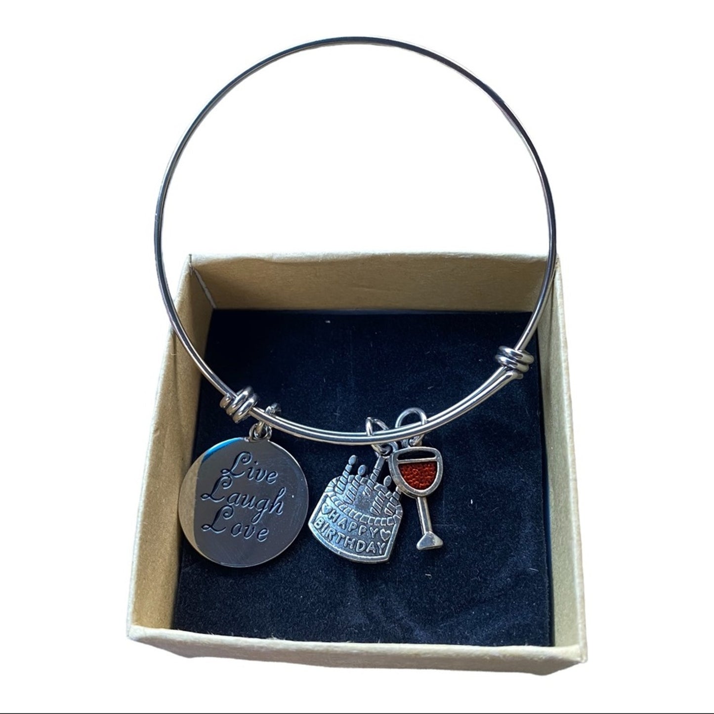 A silver charm bangle bracelet displayed inside its beige gift box. The bracelet features three charms: a round silver disc engraved with 'Live Laugh Love', a silver tiered cake charm with 'Happy Birthday' text, and a silver wine glass charm with a red liquid accent.