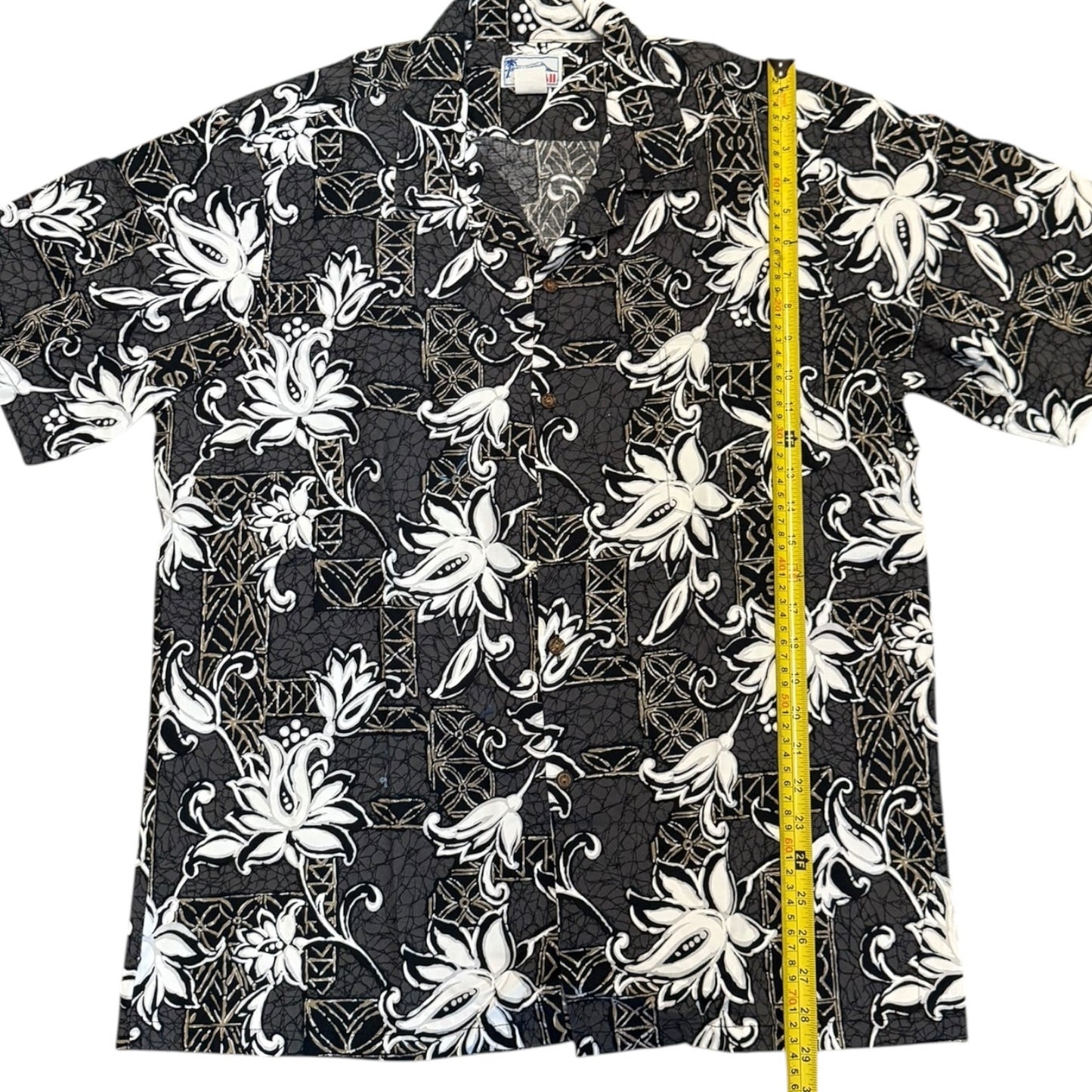 A short-sleeve button-up aloha shirt with a black, white, and gray tropical floral pattern, shown flat with a yellow tape measure standing vertically on the right side for size reference.