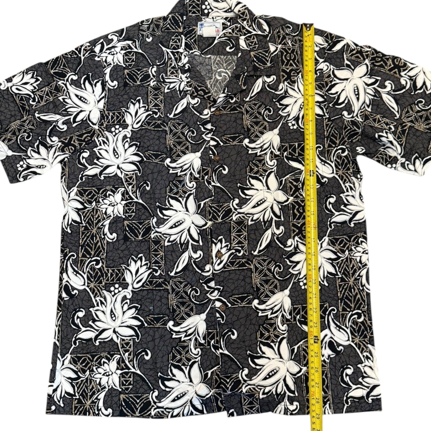 A short-sleeve button-up aloha shirt with a black, white, and gray tropical floral pattern, shown flat with a yellow tape measure standing vertically on the right side for size reference.