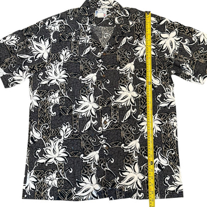 A short-sleeve button-up aloha shirt with a black, white, and gray tropical floral pattern, shown flat with a yellow tape measure standing vertically on the right side for size reference.