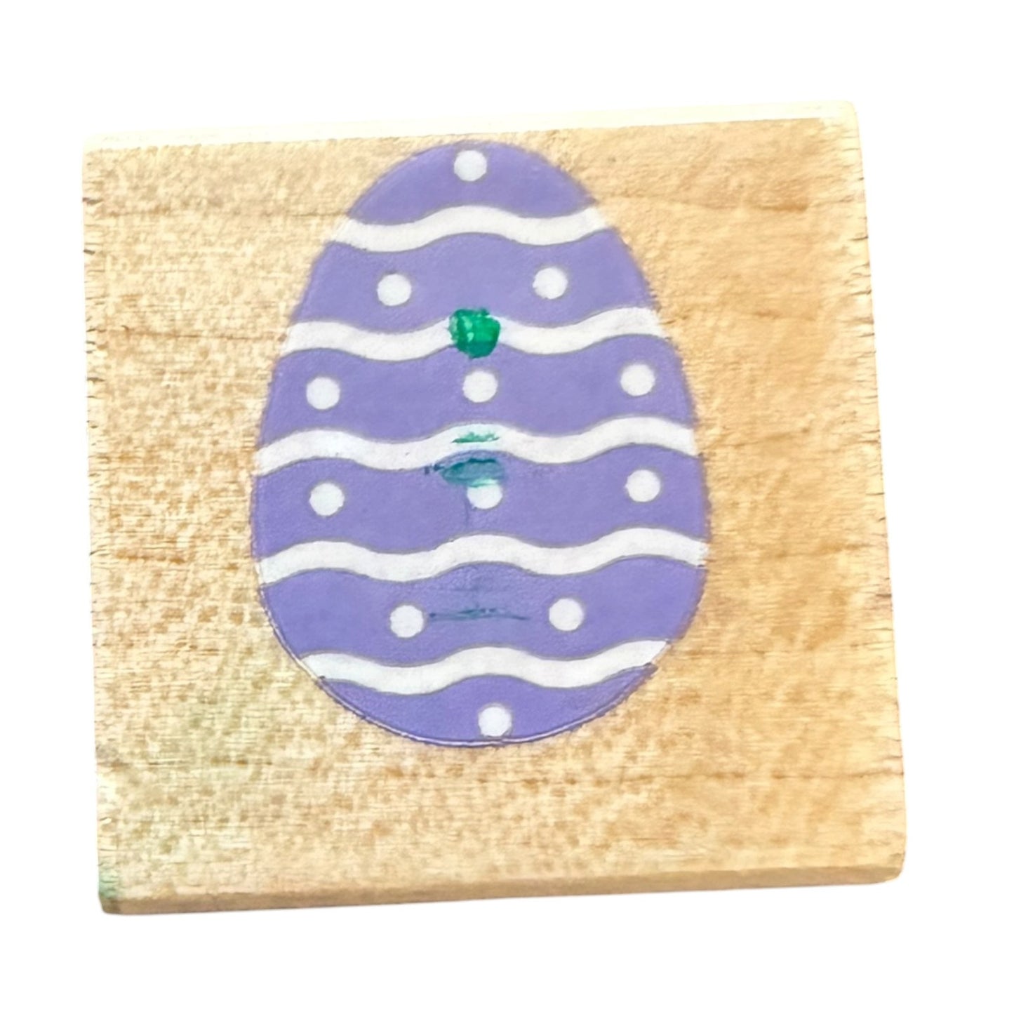 A square wooden block featuring a  purple Easter egg design with white wavy stripes, white dots, and a small green accent, shown against a plain white background.