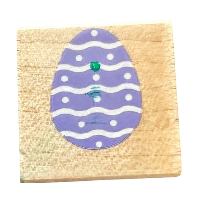 A square wooden block featuring a  purple Easter egg design with white wavy stripes, white dots, and a small green accent, shown against a plain white background.