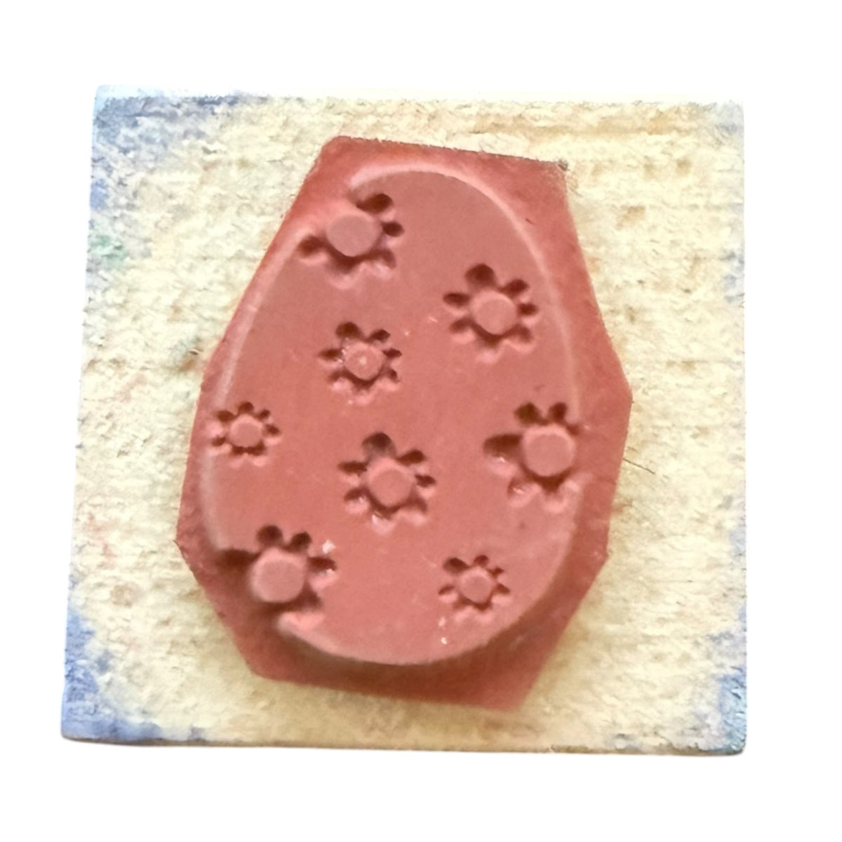 A terracotta-colored rubber stamp with a floral pattern on an egg, shown on a textured beige paper square against a white background.