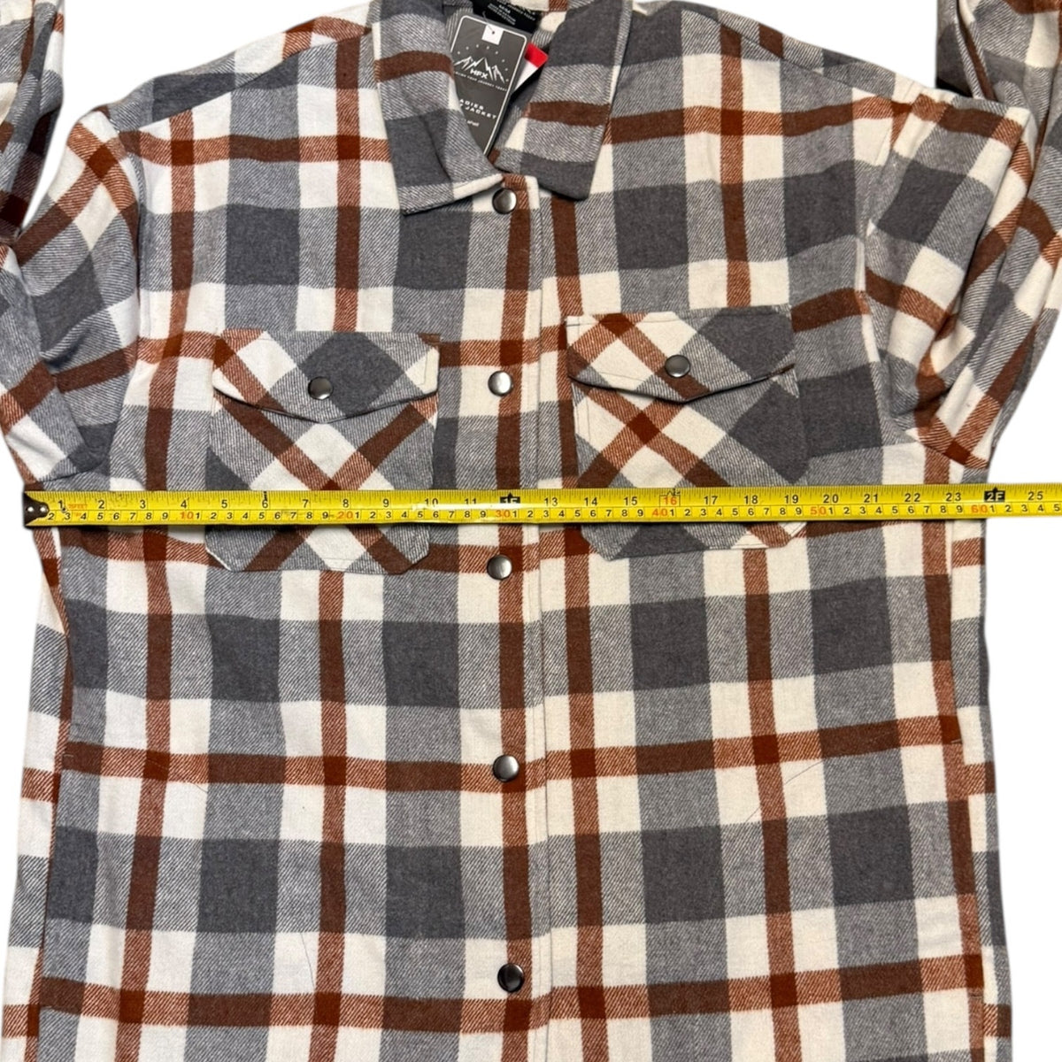 A long-sleeve plaid button-up shirt laid flat on a white background, with a yellow tape measure stretched horizontally across the chest to show the width. 