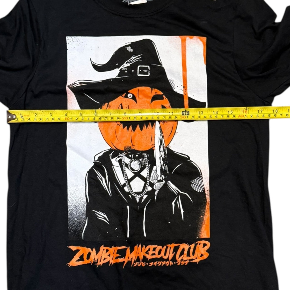 A black short-sleeve graphic T-shirt laid flat on a white background, with a yellow tape measure stretched horizontally across the chest to show the garment width. 
