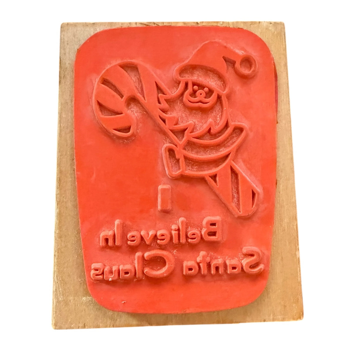 A Christmas-themed rubber stamp on a wooden block, featuring a Santa Claus holding a candy cane with the text 'Believe In Santa Claus' in reverse, shown against a plain white background.