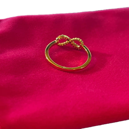 A gold ring with a twisted infinity symbol design, displayed on a vibrant magenta satin fabric background.