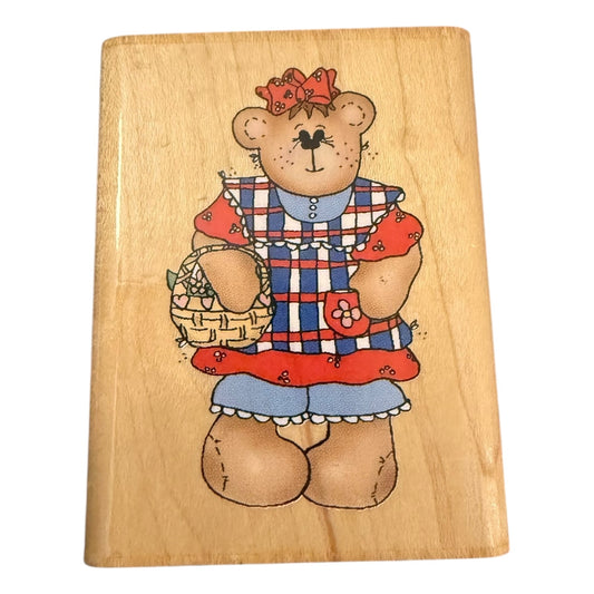 Wooden block with a teddy bear illustration wearing a dress and holding a basket.