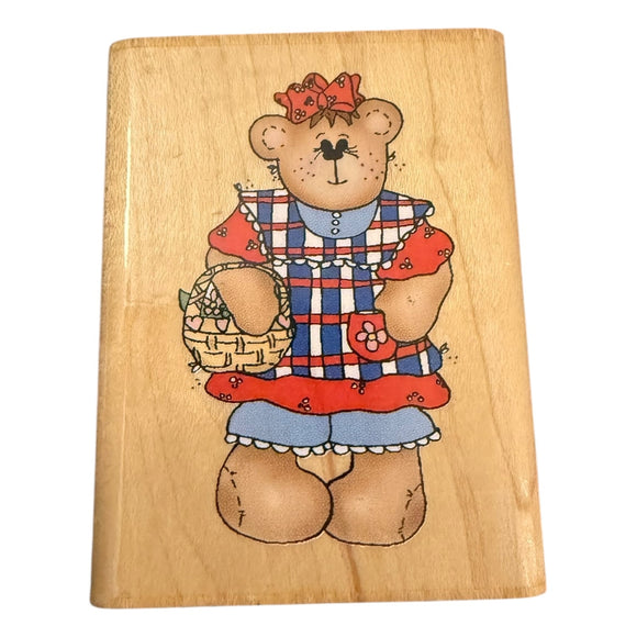 Wooden block with a teddy bear illustration wearing a dress and holding a basket.