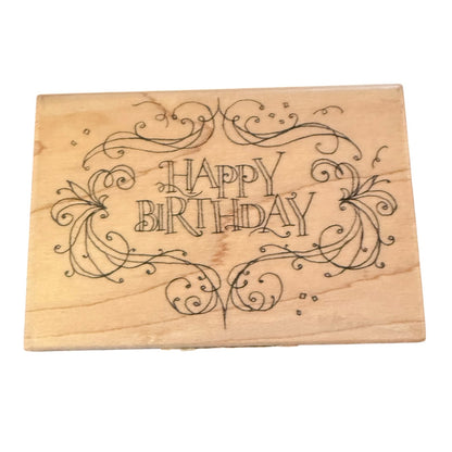 A rectangular wooden stamp featuring the words 'HAPPY BIRTHDAY' in decorative black scrollwork, shown on a plain white background.