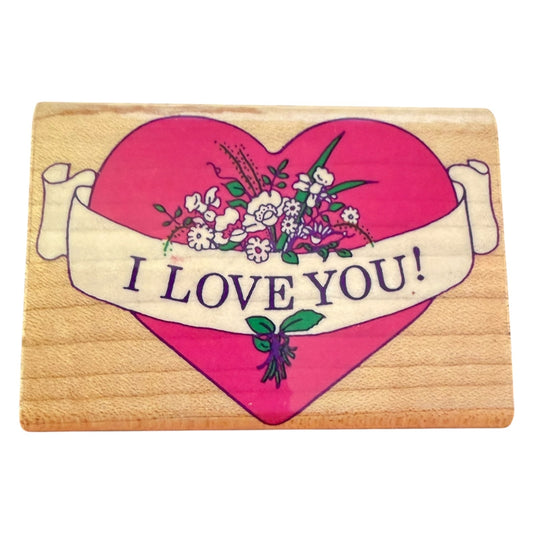 A rectangular wooden rubber stamp featuring a large pink heart with a white banner across the middle that reads “I LOVE YOU!” in dark lettering. The heart is decorated with small white, pink, and green flowers and leaves.