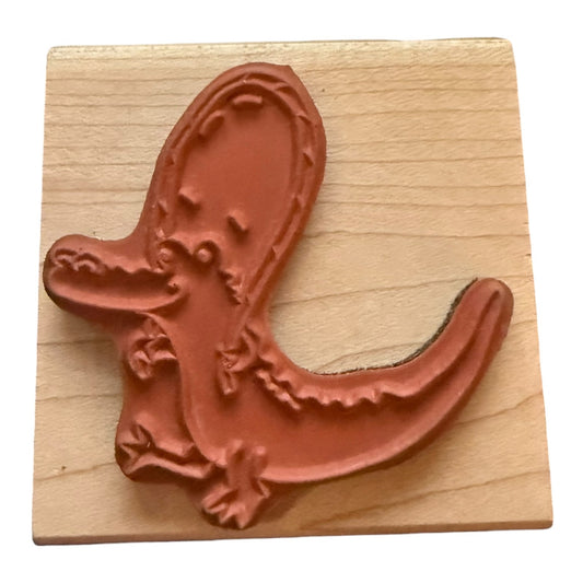 A rubber stamp featuring a stylized cartoon alligator, mounted on a square light wood block against a white background.