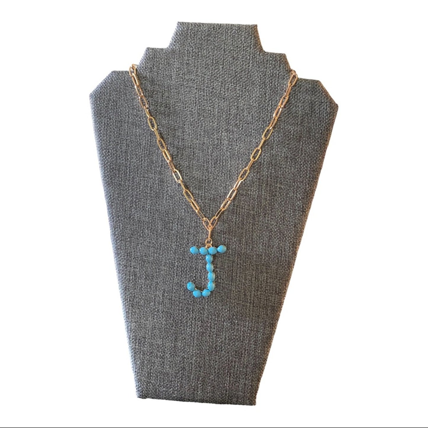 A gold chain necklace with a letter 'J' pendant made of small turquoise-blue beads, displayed on a gray fabric jewelry bust against a white background.