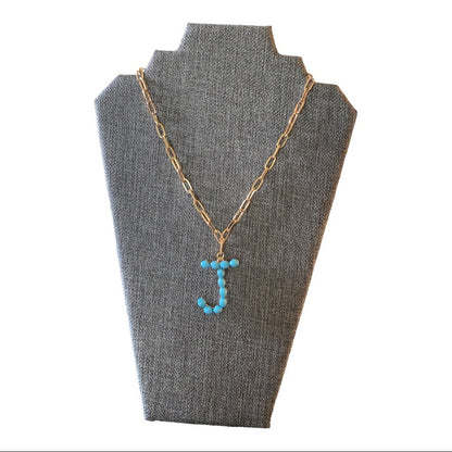 A gold chain necklace with a letter 'J' pendant made of small turquoise-blue beads, displayed on a gray fabric jewelry bust against a white background.