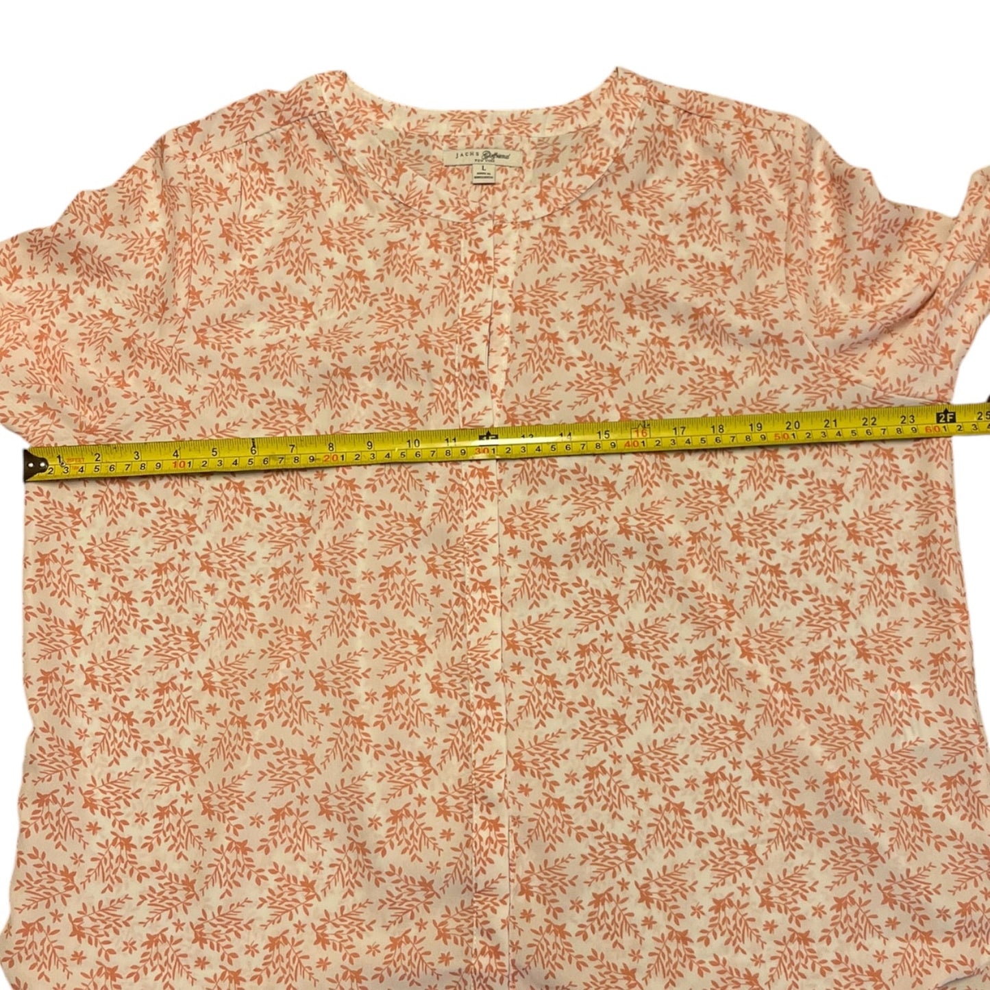 A button-up shirt with a pink floral pattern on a white background, shown flat with a yellow tape measure stretched horizontally across the waist to indicate the garment's width.