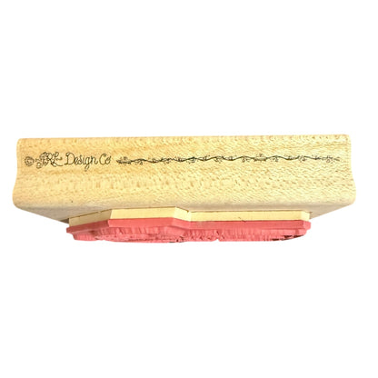 A close-up of a stamp block with a textured beige wooden top featuring the brand name 'JRL Design Co' in black script, supported by layered pink and beige rubber stamps, shown against a plain white background.