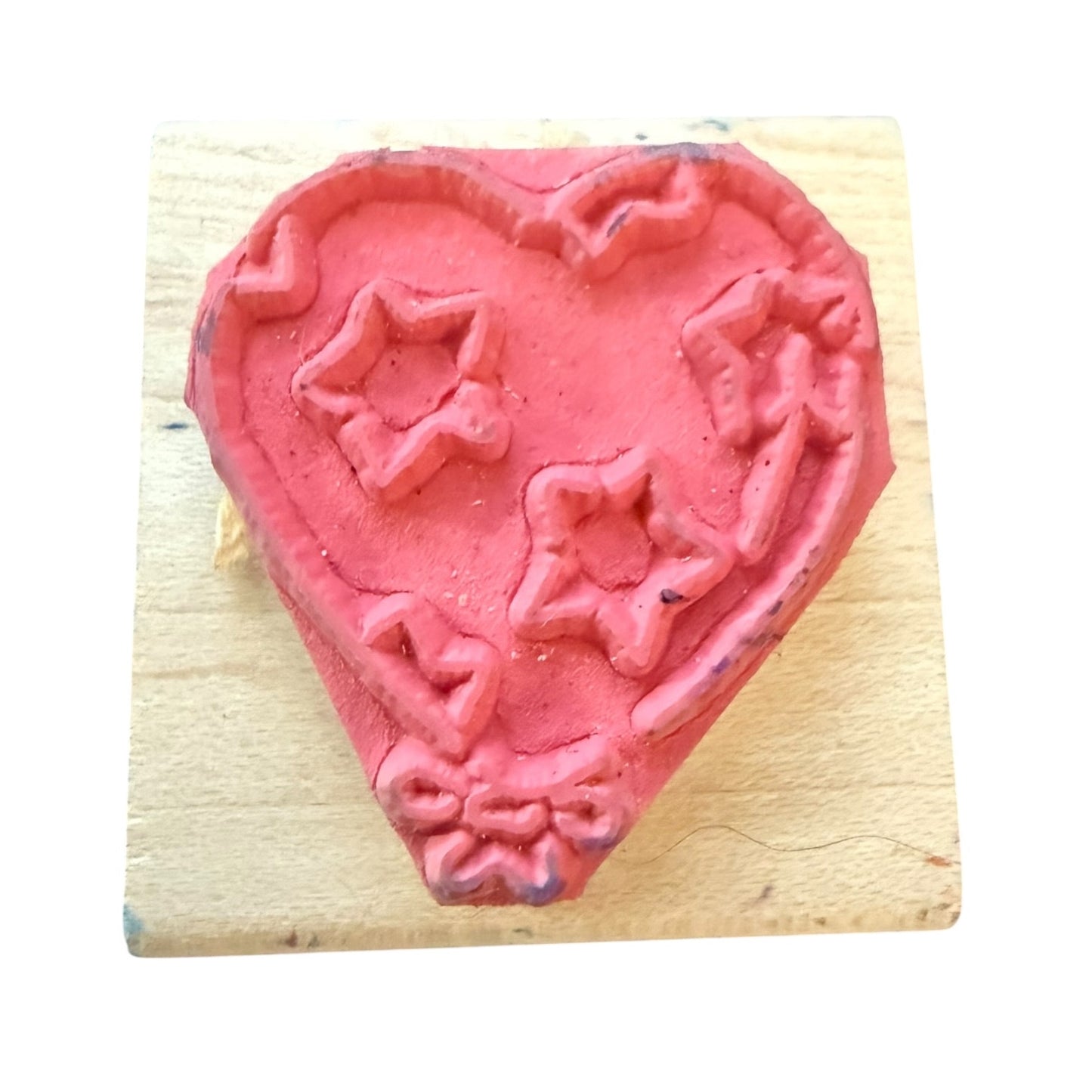 A pink heart-shaped rubber stamp with embossed star and bow patterns, resting on a light-colored wooden block against a white background.