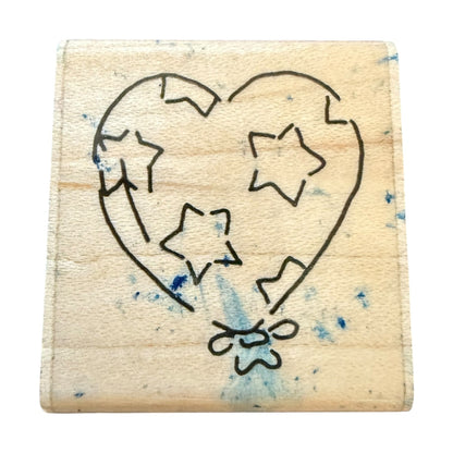 A square wooden rubber stamp block featuring a heart balloon and star design with blue ink splatters, shown on a plain white background.