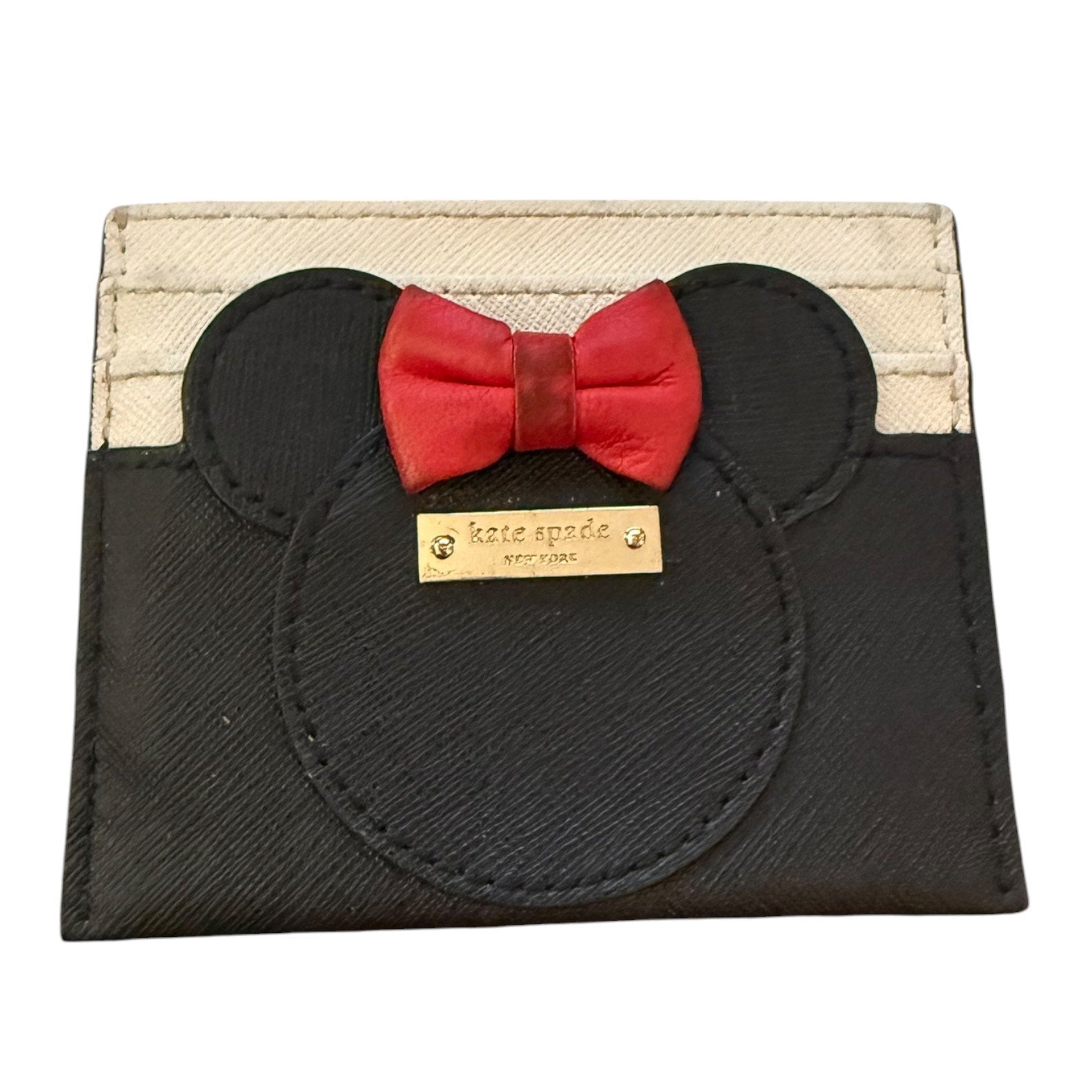 A Kate Spade New York card holder featuring a black Minnie Mouse silhouette with a red bow tie and a gold-tone brand plaque, set against a cream canvas background.
