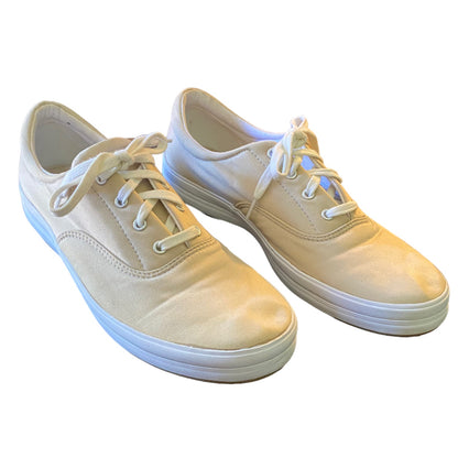 A pair of low-top canvas sneakers in a beige color, featuring white laces and white rubber soles, shown against a plain white background.