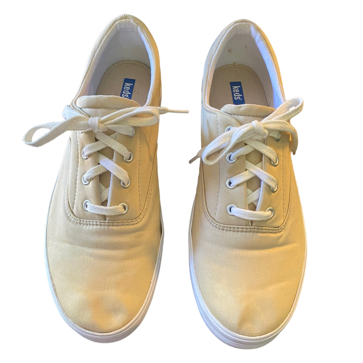 A pair of canvas low-top sneakers in a beige color with white laces, shown from a top-down view on a white background.