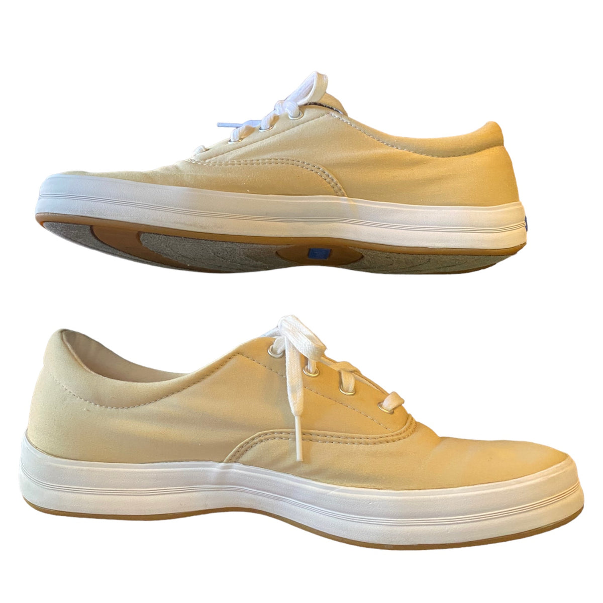 A pair of low-top canvas sneakers shown from the side, featuring a light beige canvas upper, white laces, a thick white midsole, and a brown gum outsole, set against a plain white background.