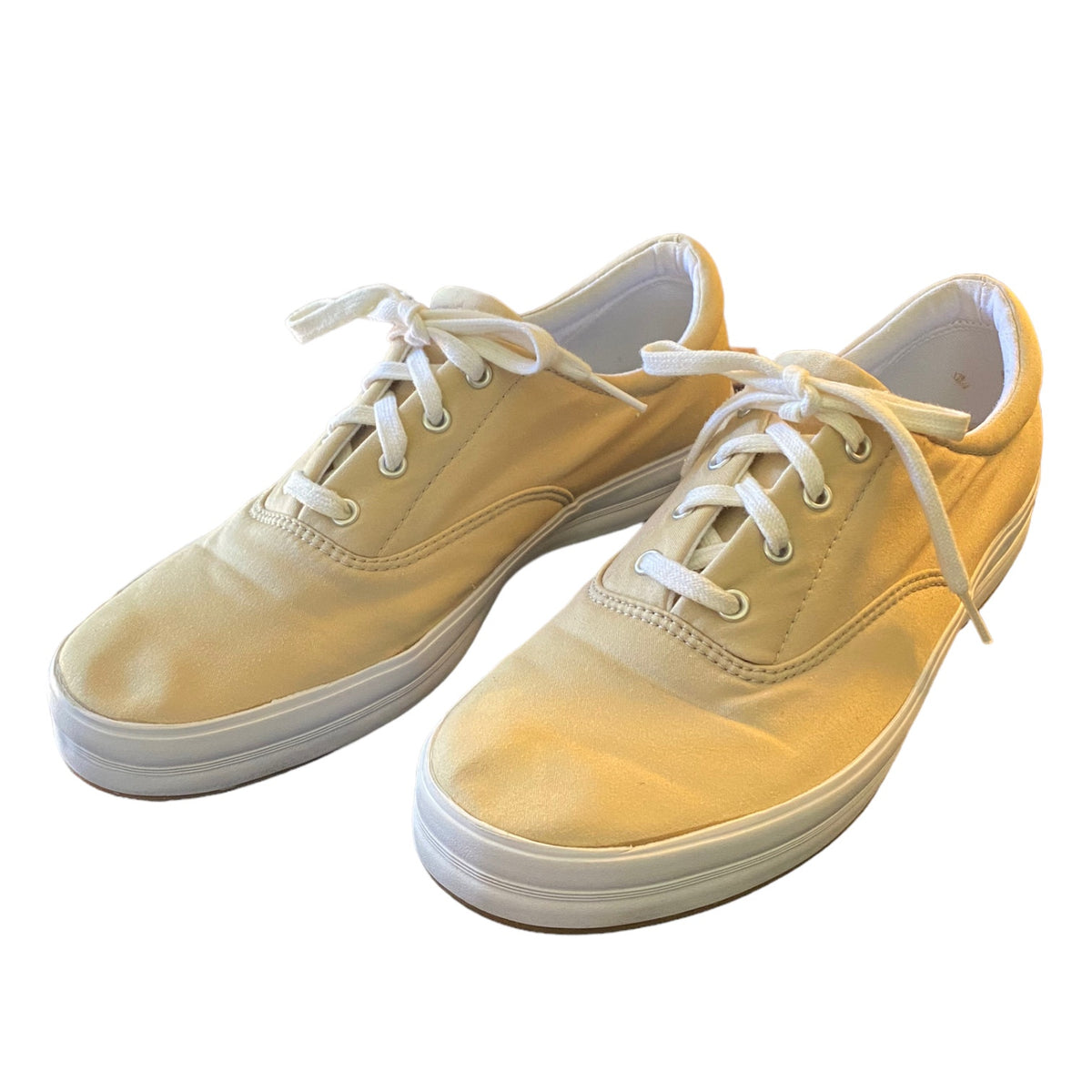 A pair of low-top canvas sneakers in a beige color, featuring white laces and white rubber soles, set against a plain white background.