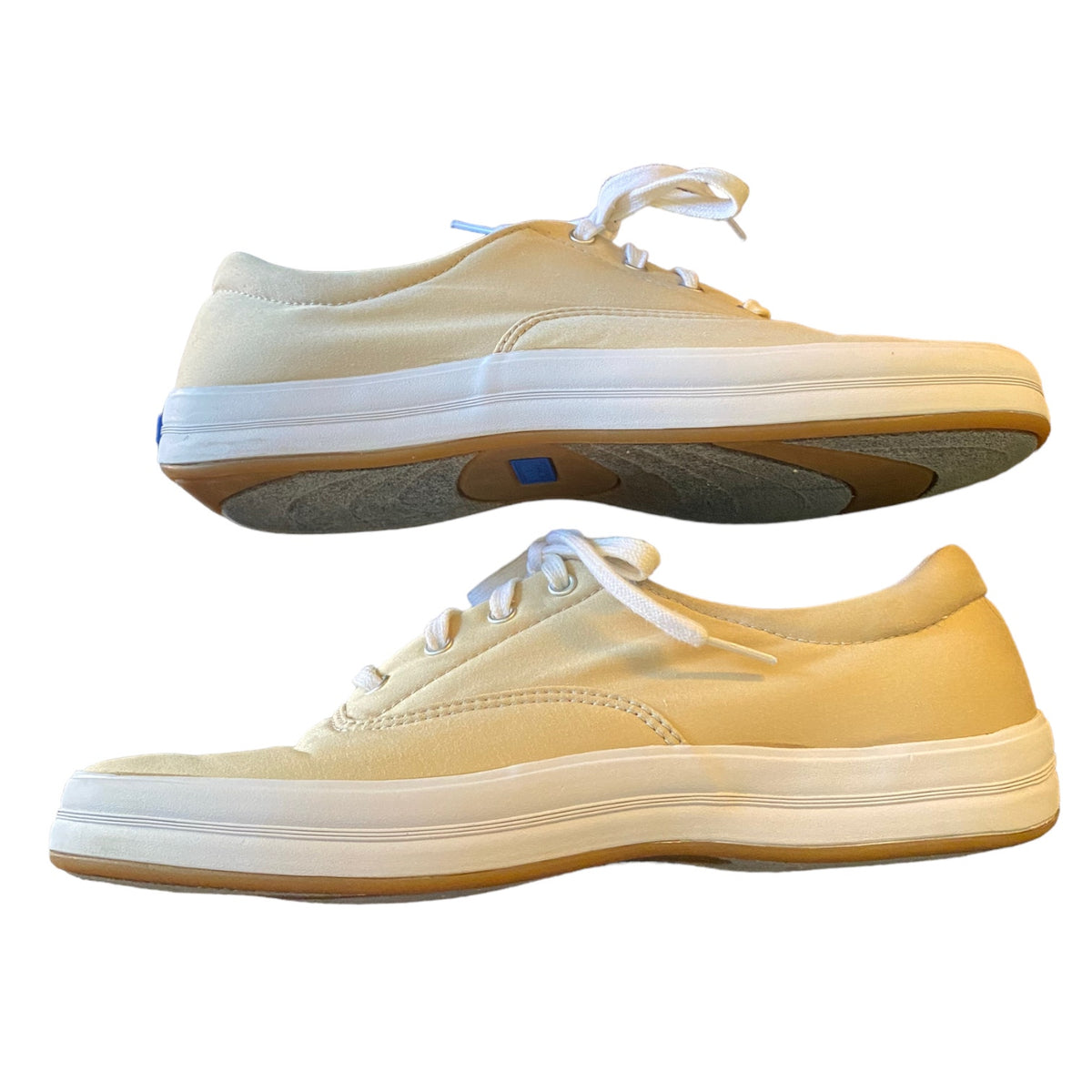 A pair of low-top canvas sneakers in a beige color, shown from the side against a plain white background. One shoe is positioned above the other, displaying the side profile, laces, and gum rubber sole.
